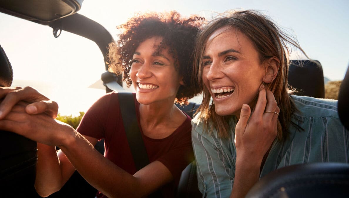 Nahaufnahme von zwei jungen, lachenden Frauen mit wehenden Haaren auf dem Rücksitz eines Cabriolets.