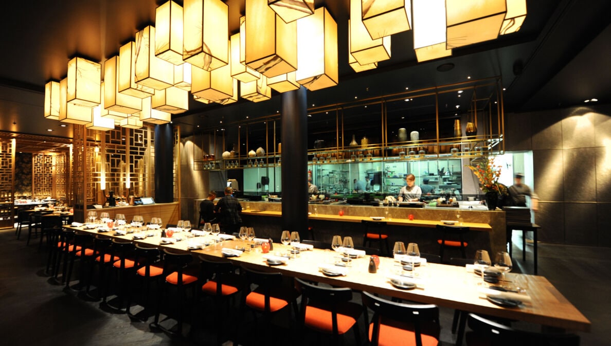 Ein langer, eingedeckter Tisch in einem modernen Restaurant im fernöstlichen Stil mit offener Küche bei gedimmter Beleuchtung.