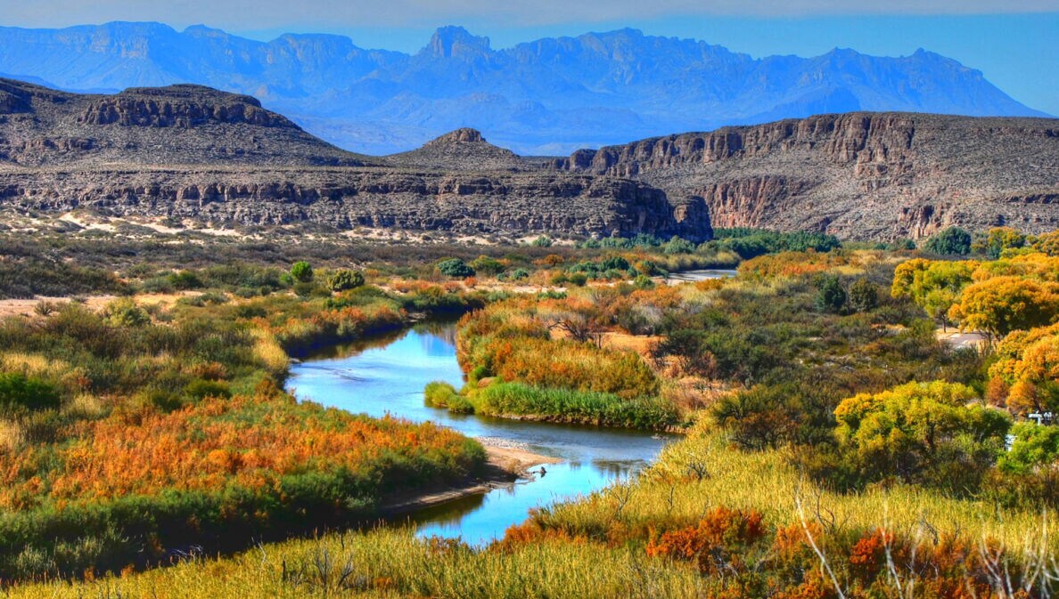 Landschaftspanorama im Big-Bend-Nationalpark mit mäanderndem Fluss in einer herbstlich gefärbten Graslandschaft vor zwei Gebirgsketten.