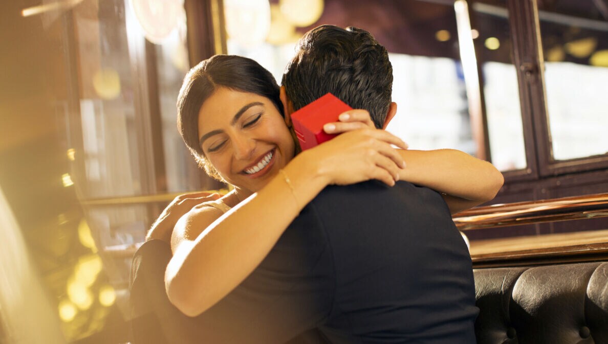 Eine Frau mit einer roten Geschenkbox in den Händen umarmt glücklich lächelnd einen Mann auf einer Sitzbank in einem Restaurant.