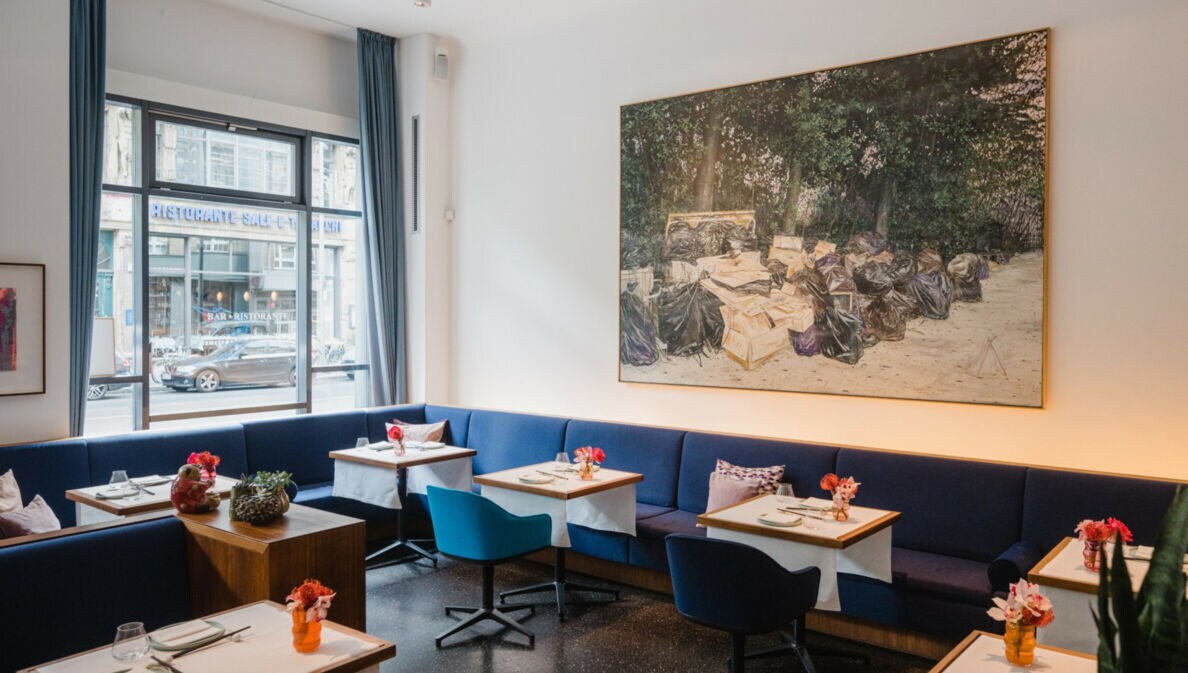 Innenraum eines hellen, modernen Restaurants mit blauen Sitzmöbeln an gedeckten Tischen und einem großen Gemälde an der Wand.