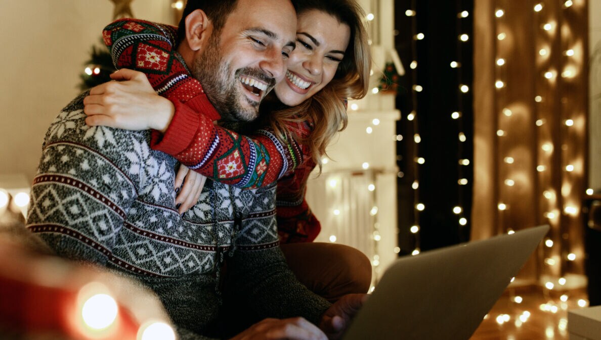 Ein lachendes Paar in Winterpullovern sitzt aneinander gekuschelt auf einem Sofa im Wohnzimmer vor einem Laptop.