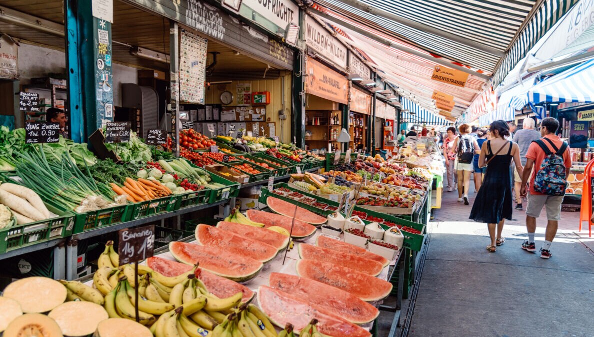 Personen auf einer Marktstraße mit Obst- und Gemüseauslagen.