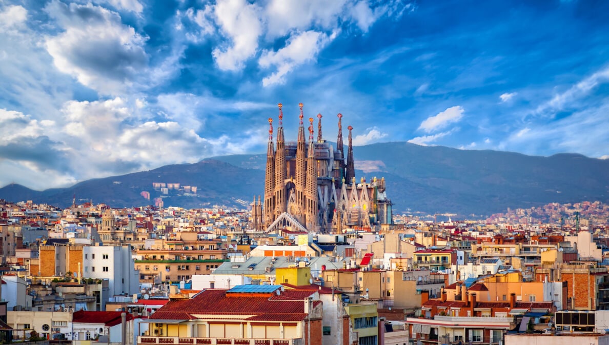 Stadtpanorama von Barcelona mit der emporragenden Sagrada Familia vor einem Gebirgszug im Hintergrund.