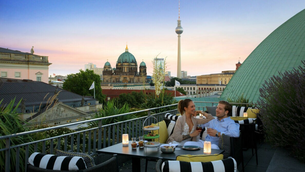 Ein junges Paar sitzt gut gelaunt an einem Tisch auf einer Dachterrasse, im Hintergrund Berliner Dom und Fernsehturm.