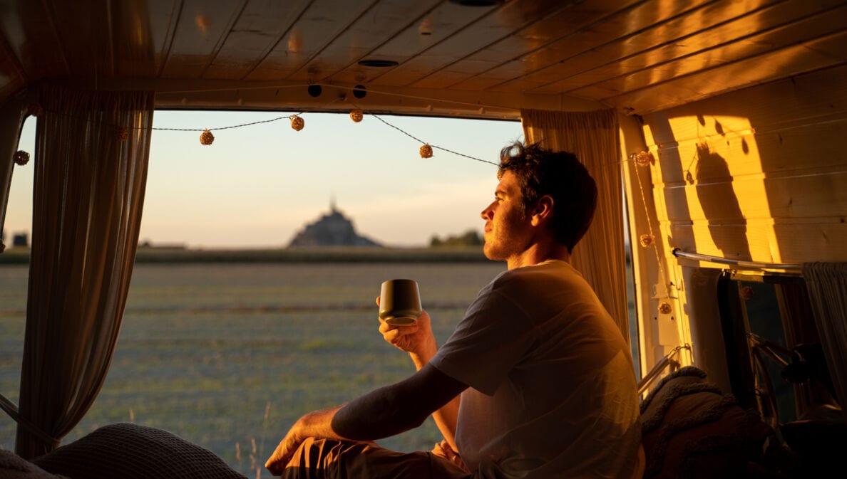 Ein junger Mann sitzt mit einem Kaffeebecher in der Hand im geöffneten Campervan im goldenen Sonnenlicht und blickt hinaus in die Natur.