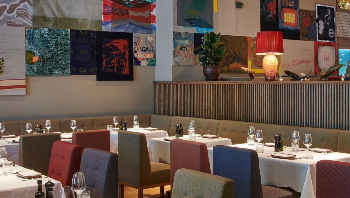 Restaurant mit Polsterstühlen an elegant gedeckten Tischen vor einer Wand mit farbenfrohen Gemälden.