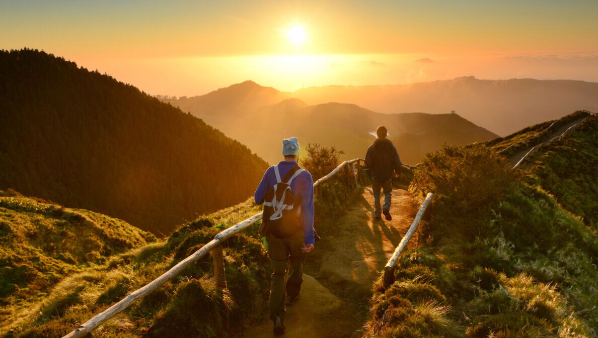 Rückansicht zweier gehender Personen auf einem Weg entlang eines Bergkamms inmitten einer grünen Gebirgslandschaft bei Sonnenuntergang.
