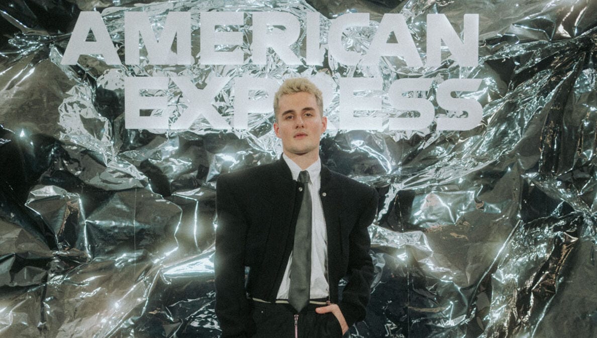 Luis Freitag von den Elevator Boys im schwarzem Anzug mit weißem Hemd und Krawatte vor einer silbernen Wand mit weißem American Express Schriftzug.