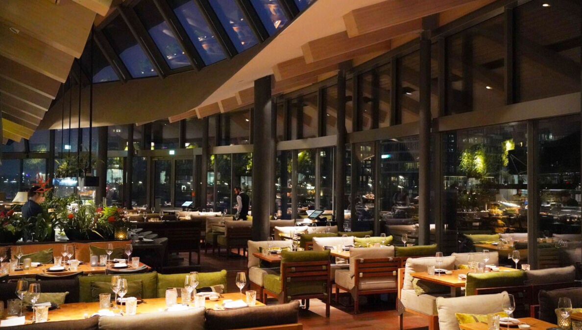 Speiseraum eines modernen Restaurants mit geschwungenem Dach und umlaufender Glasfassade bei Nacht.