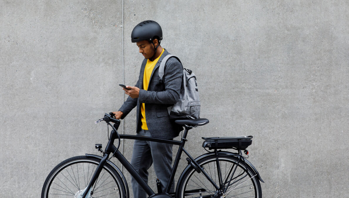 Ein junger Mann mit Rucksack und Fahrradhelm steht neben seinem E-Bike und schaut auf sein Smartphone.