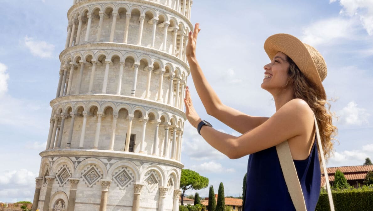 Profilansicht einer jungen Frau mit Strohhut, die aus der fotografischen Perspektive den Schiefen Turm von Pisa mit ihren Händen zu halten scheint.