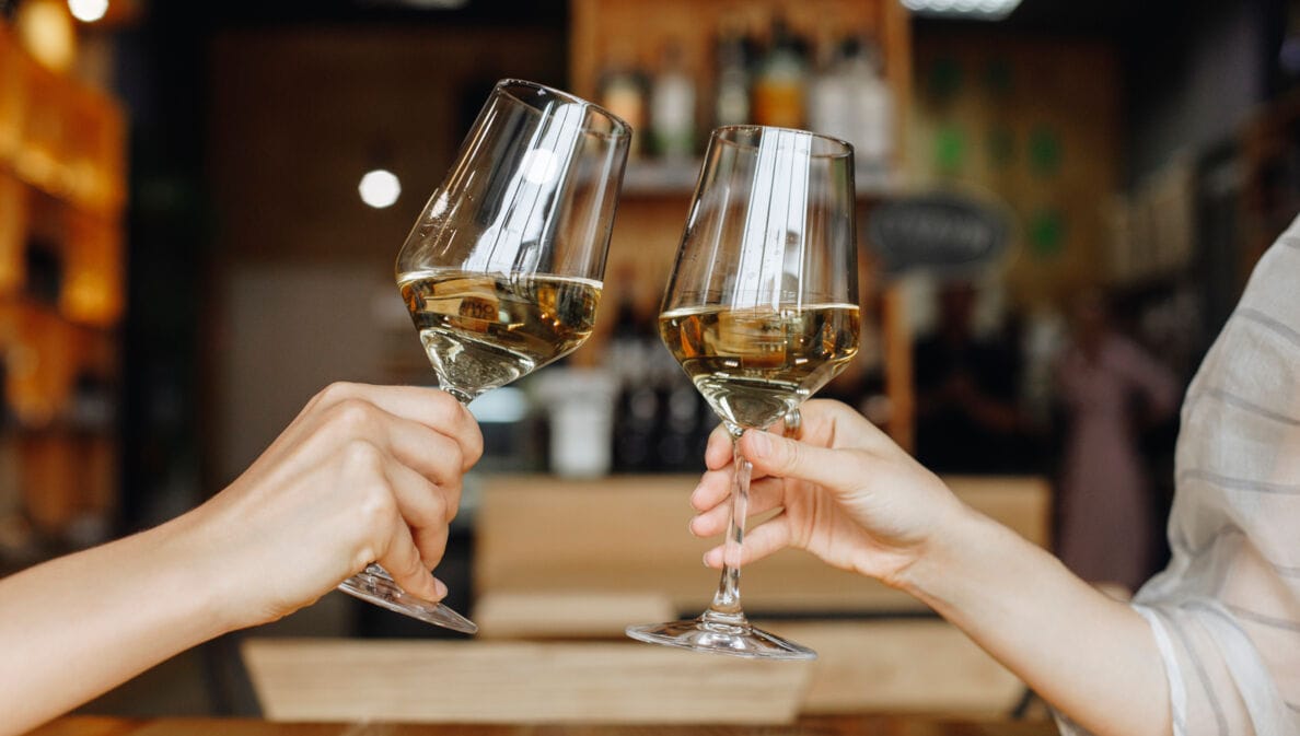 Nahaufnahme von Händen und Unterarmen von zwei Personen, die jeweils ein gefülltes Weinglas halten und anstoßen.