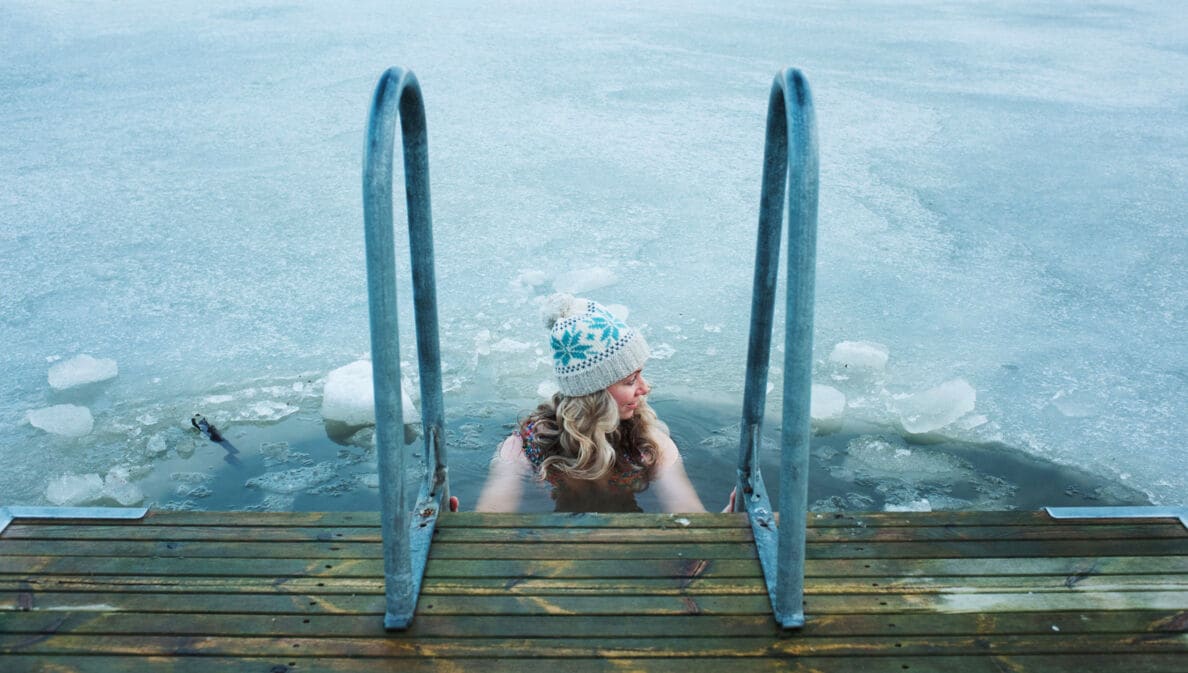 Eine Frau mit Wollmütze hält sich beim Baden in einem zugefrorenen See an dem Handlauf einer Treppe an einem Holzsteg fest.
