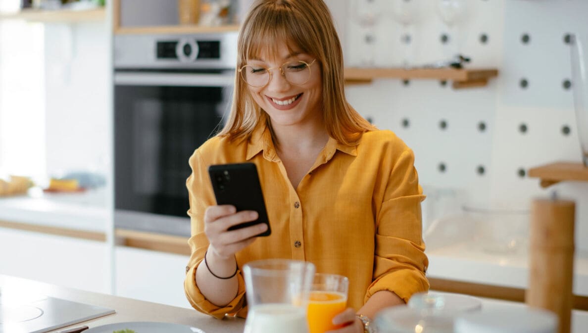 Eine junge Frau in gelbem Hemd steht mit einem Glas Orangensaft in der Hand in der Küche und schaut lächelnd auf ihr Smartphone.