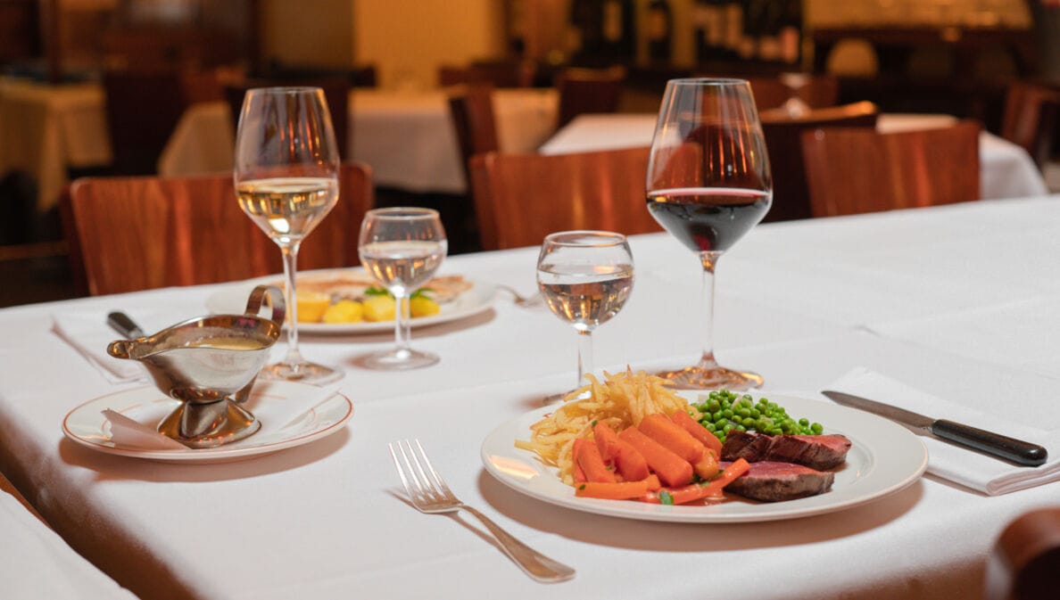 Zwei auf weißen Tellern angerichtete Speisen stehen neben Weingläsern auf einem Tisch mit weißer Stofftischdecke in einer Brasserie.
