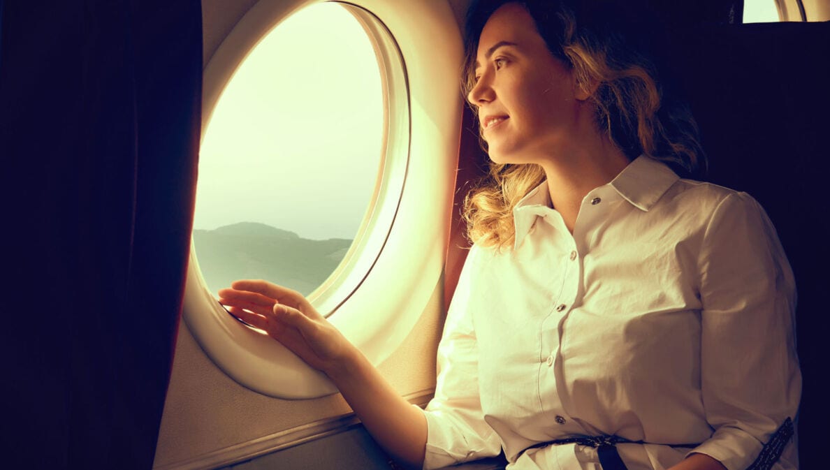 Eine junge Frau in weißer Bluse schaut aus dem Fenster eines Flugzeuges auf eine Landschaft.