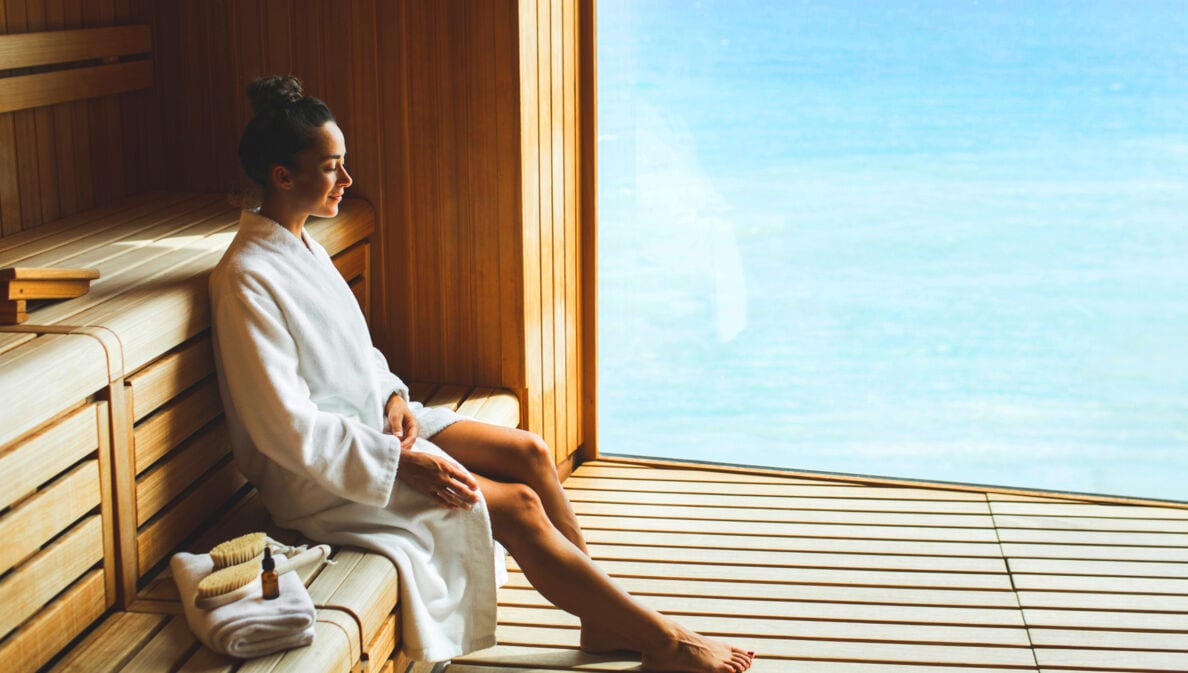 Eine junge Frau im weißen Bademantel sitzt entspannt in einer Holzsauna mit Panoramafenster am Meer.