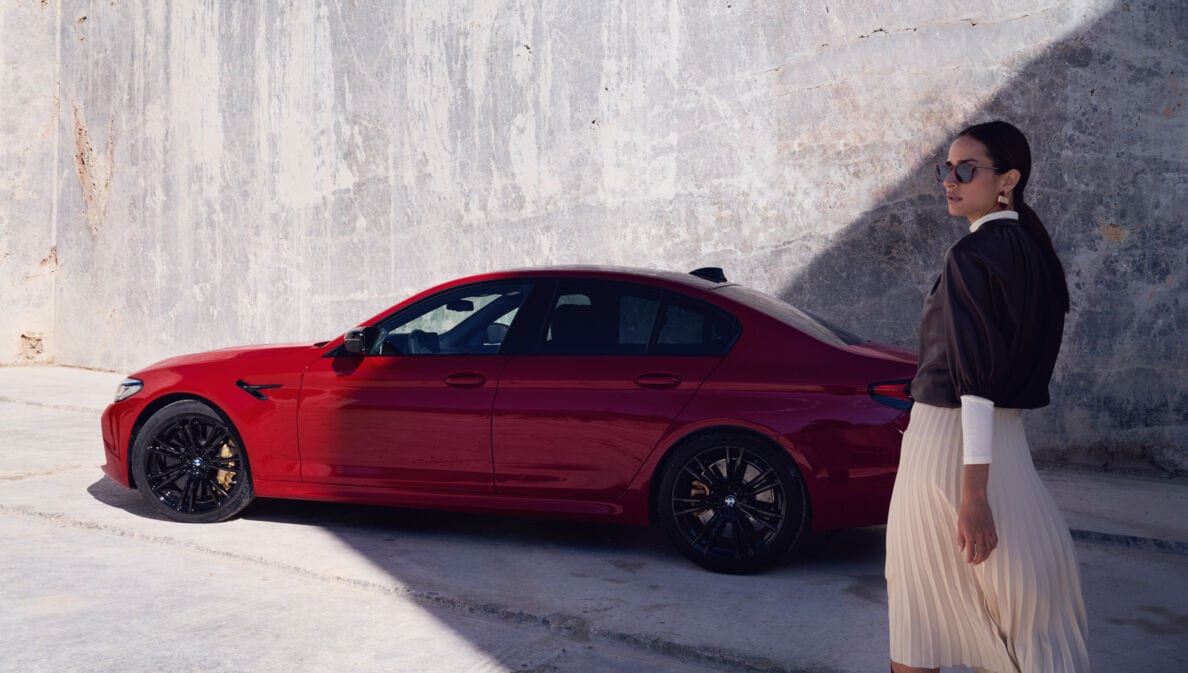 Eine elegante Frau mit Sonnenbrille vor einem roten BMW vor einer Betonwand.