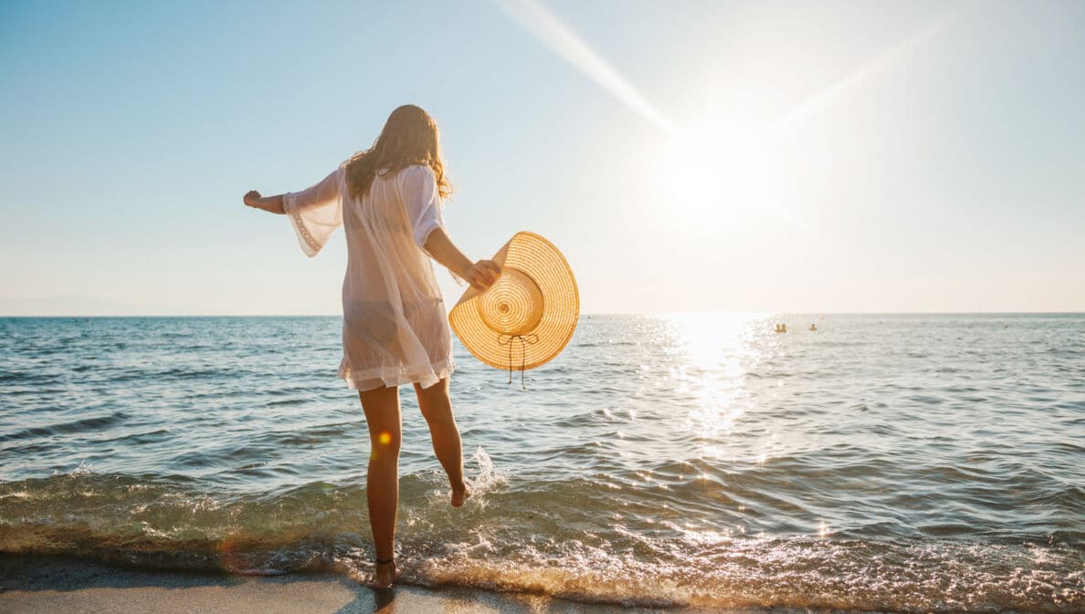 Eine junge Frau im weißen Sommerkleid und mit Strohhut in der Hand planscht mit ihrem Fuß im Wasser an einem Sandstrand am Meer bei Sonnenuntergang