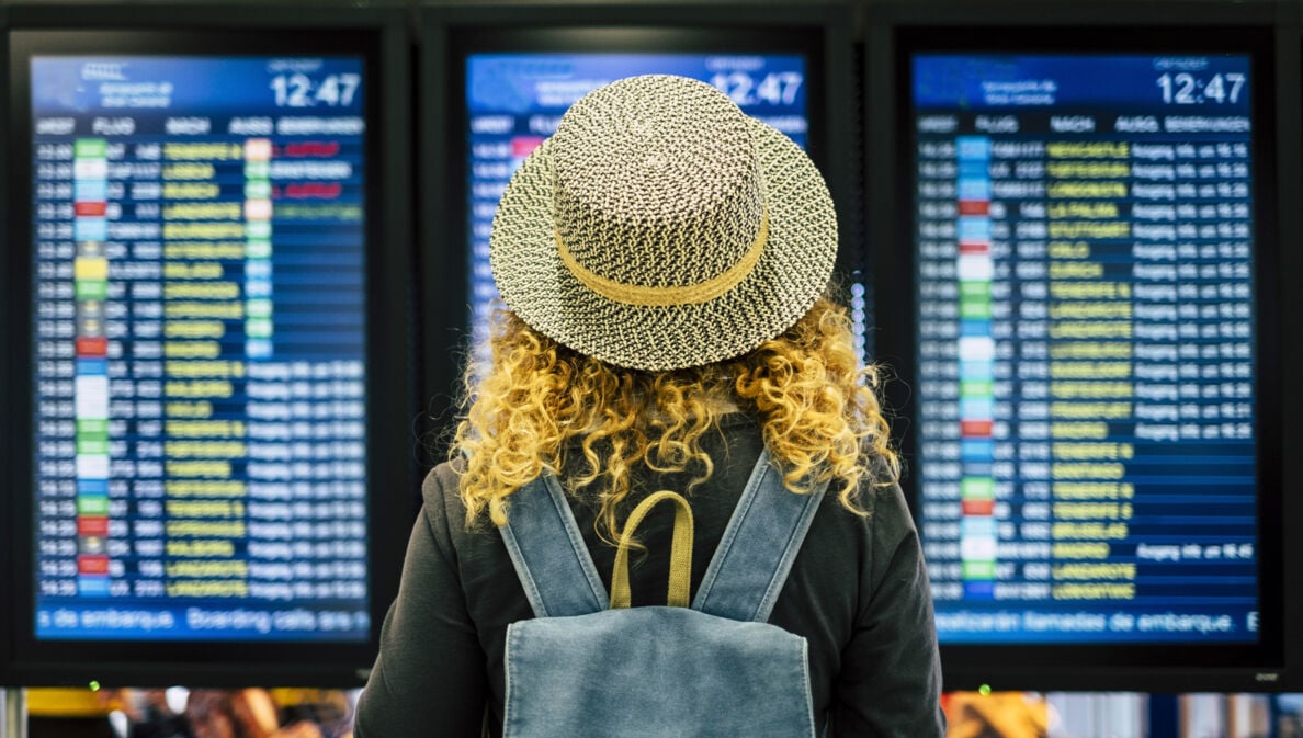 Rückansicht einer Frau mit Strohhut vor einer Anzeigetafel am Flughafen.