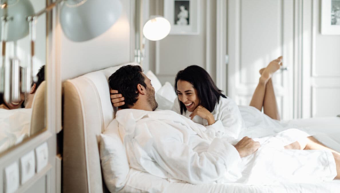 Ein Paar in weißen Bademänteln liegt lachend in einem weißen Bett in einem edlen, hellen Zimmer.