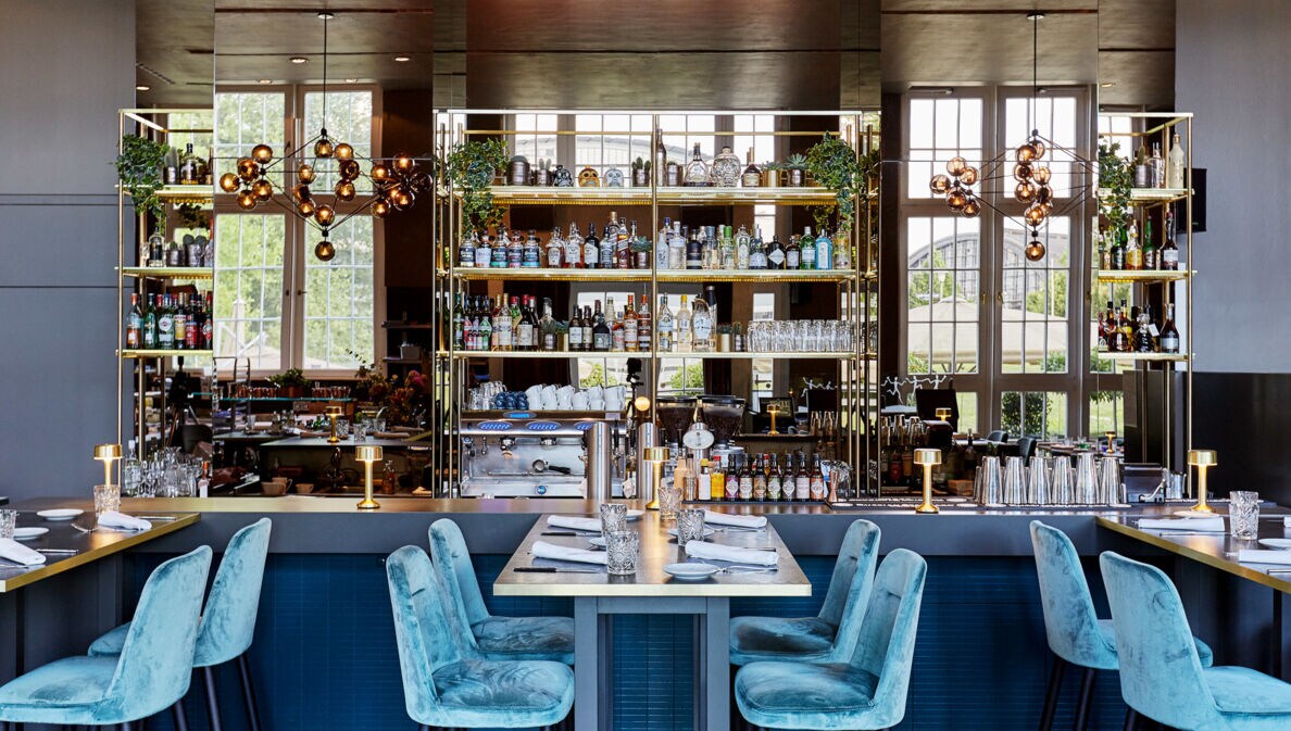 Verspiegelter Barbereich in einem modernen, edlen Restaurant mit blauen Samtstühlen an eingedeckten Tischen.