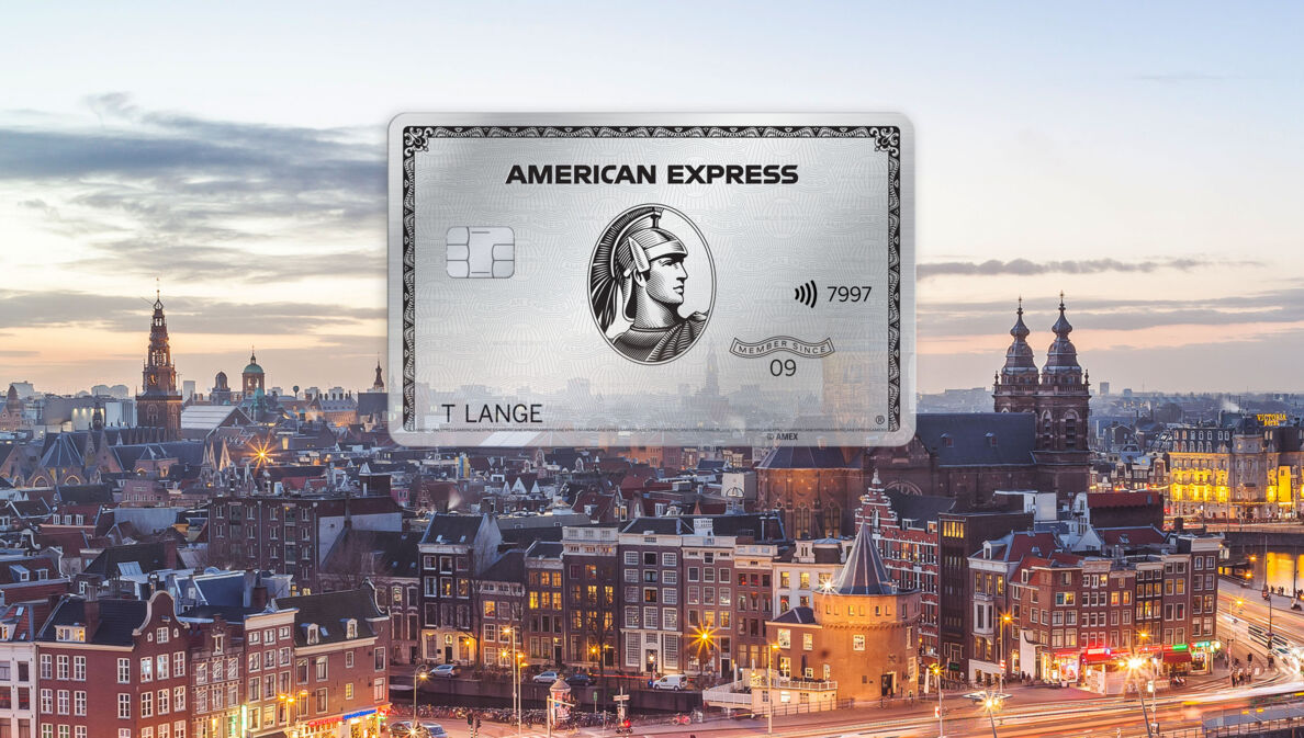 Fotocollage mit Freisteller einer silbernen Kreditkarte von American Express vor dem Stadtpanorama von Amsterdam in der Abenddämmerung.