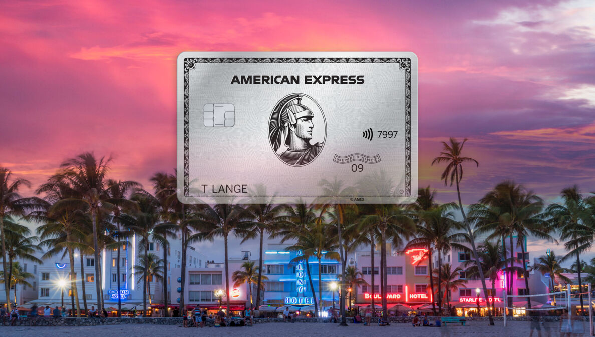 Freisteller einer Platinum Card von American Express vor der erleuchteten Oceanfront von Miami Beach hinter Palmen bei Sonnenuntergang.