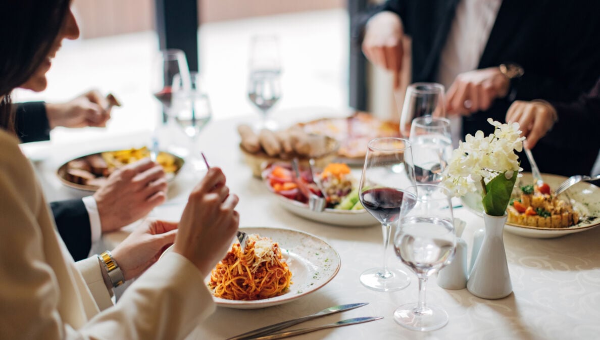 Eine Gruppe Personen an einem weiß eingedeckten Tisch in einem Restaurant mit verschiedenen Gerichten wie beispielsweise Pasta.