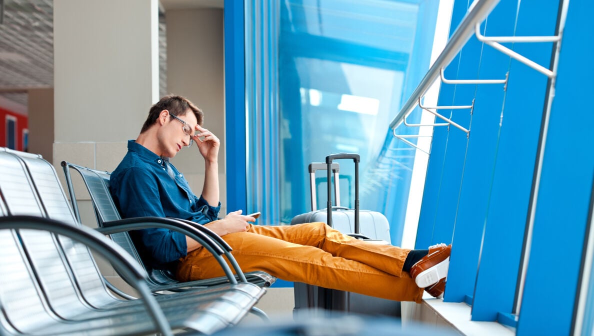 Ein Mann, der am Flughafen auf einer Bankreihe mit ausgestreckten Beinen sitzt. In einer Hand hält er ein Smartphone, die andere Hand ist an seiner Schläfe. Neben ihm steht ein Koffer.
