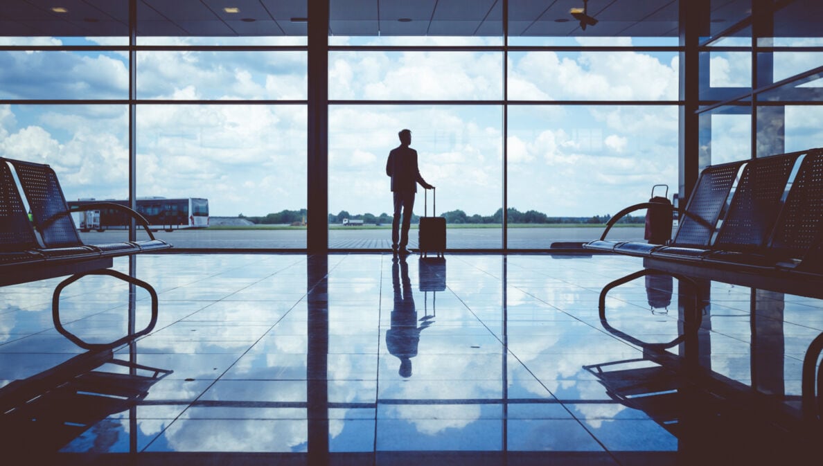 Rückenansicht einer Person, die in einer Flughafenhalle mit großer Glasfront am Fenster steht und einen Rollkoffer in einer Hand am Griff hält.