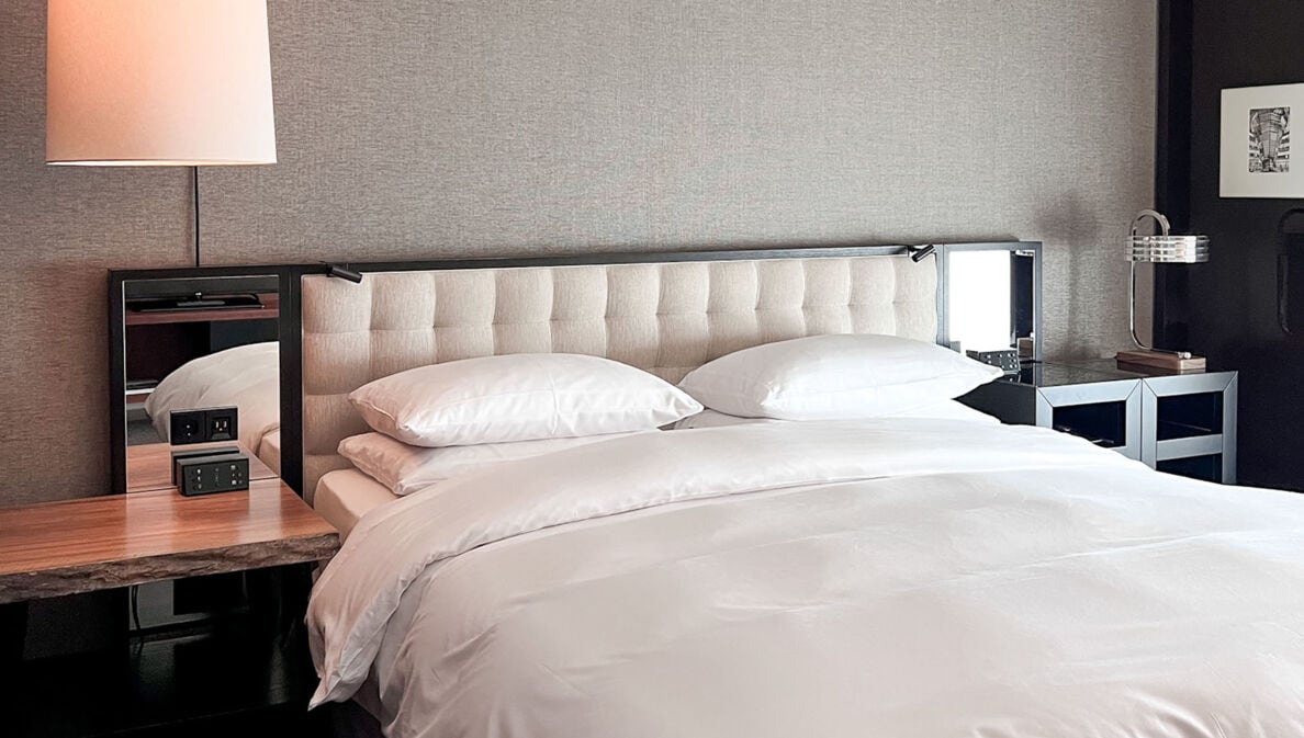 Doppelbett mit gepolstertem Kopfteil und Spiegeln sowie Nachttischen im modernen, minimalistischen Design vor einer grau tapezierten Wand.