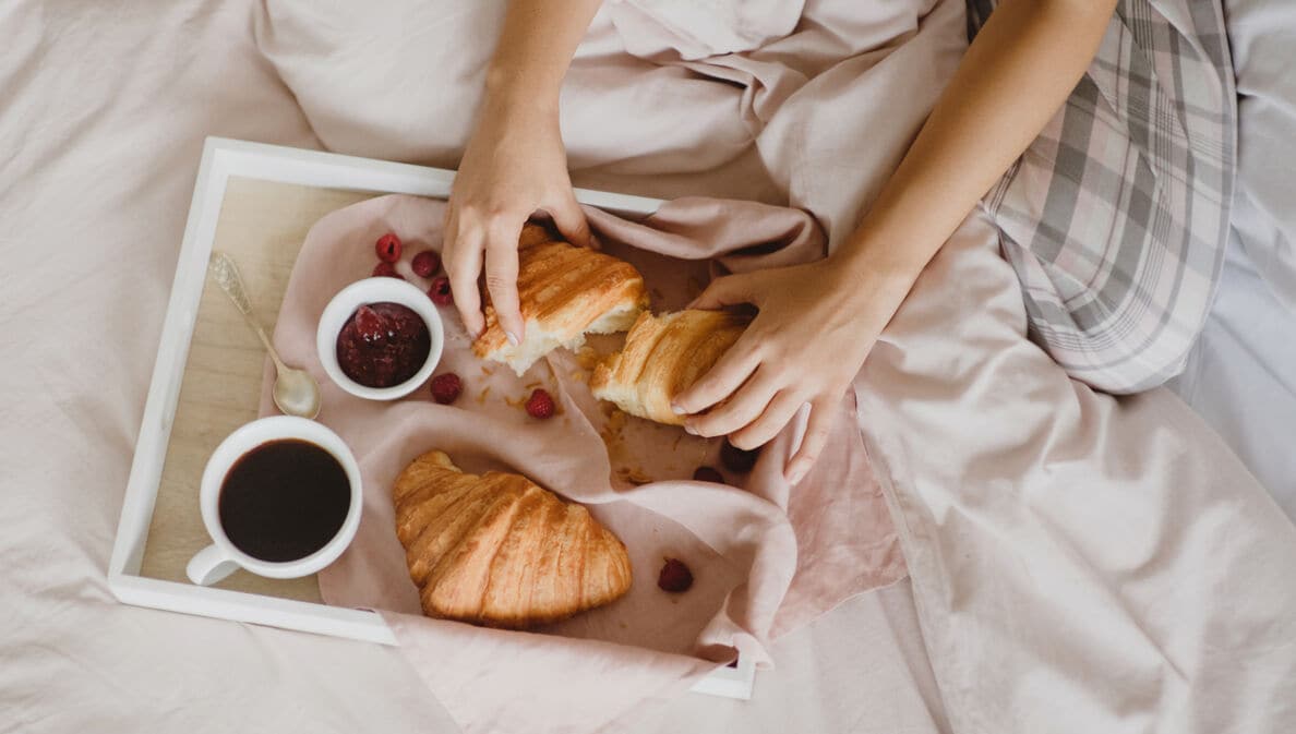 Auf einem Bett liegt ein Tablett mit Croissants, einer Tasse Kaffee, ein Schälchen Marmelade, einem Löffel und Früchten. Dahinter sitzt eine unkenntliche Person, die eines der Croissants auseinanderreißt.