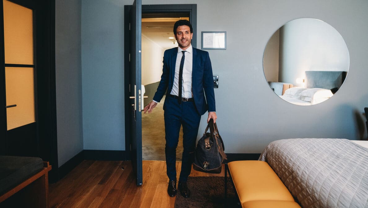 Ein Mann im Anzug trägt eine Reisetasche und betritt ein Hotelzimmer. 