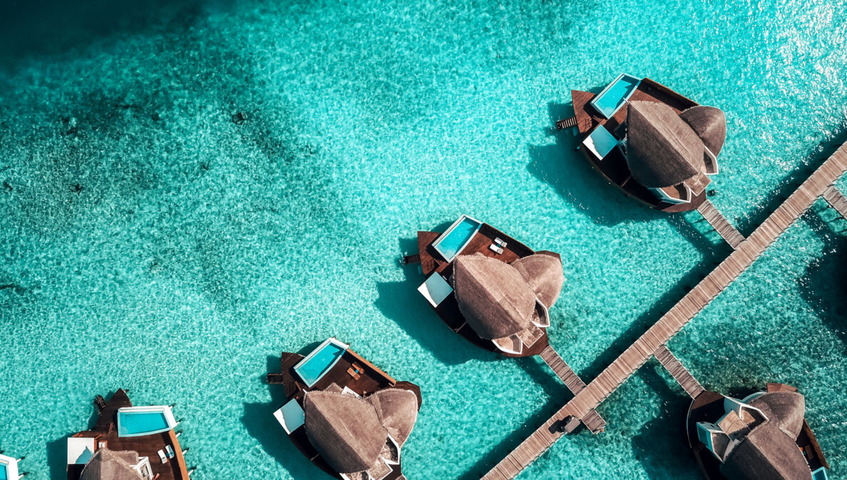 Eine Luftaufnahme einer Hotelanlage mit privaten Suiten samt Pool auf den Malediven, die in einer Linie an einem Steg angelegt und von türkisblauem Wasser umgeben sind