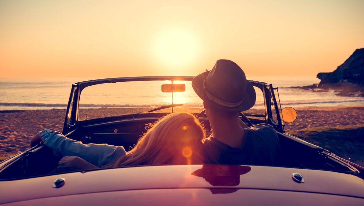 Rückansicht eines Paares in einem Cabriolet an einem Sandstrand bei Sonnenuntergang, sie hat den Kopf auf seine Schulter gelehnt.