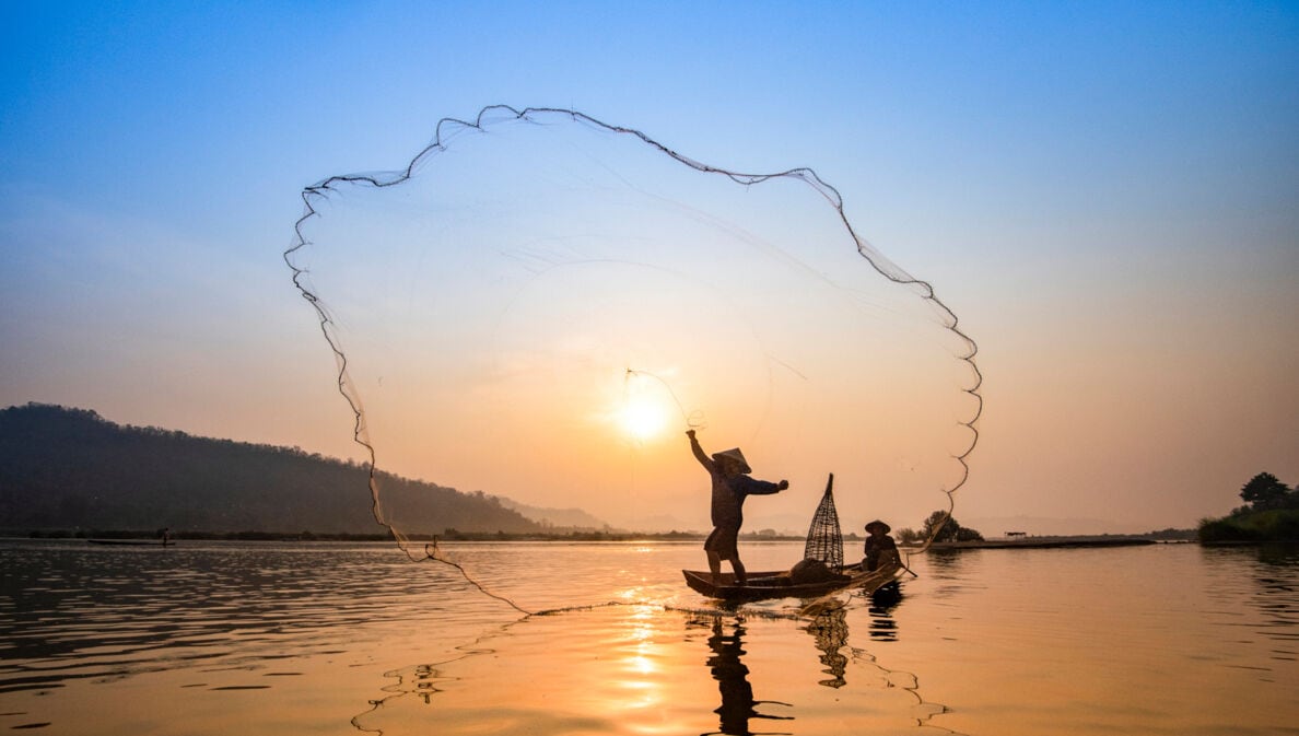 Fischer in einem Holzboot auf dem Fluss Mekong, der bei Sonnenauf- oder Untergang sein Netz auswirft.  