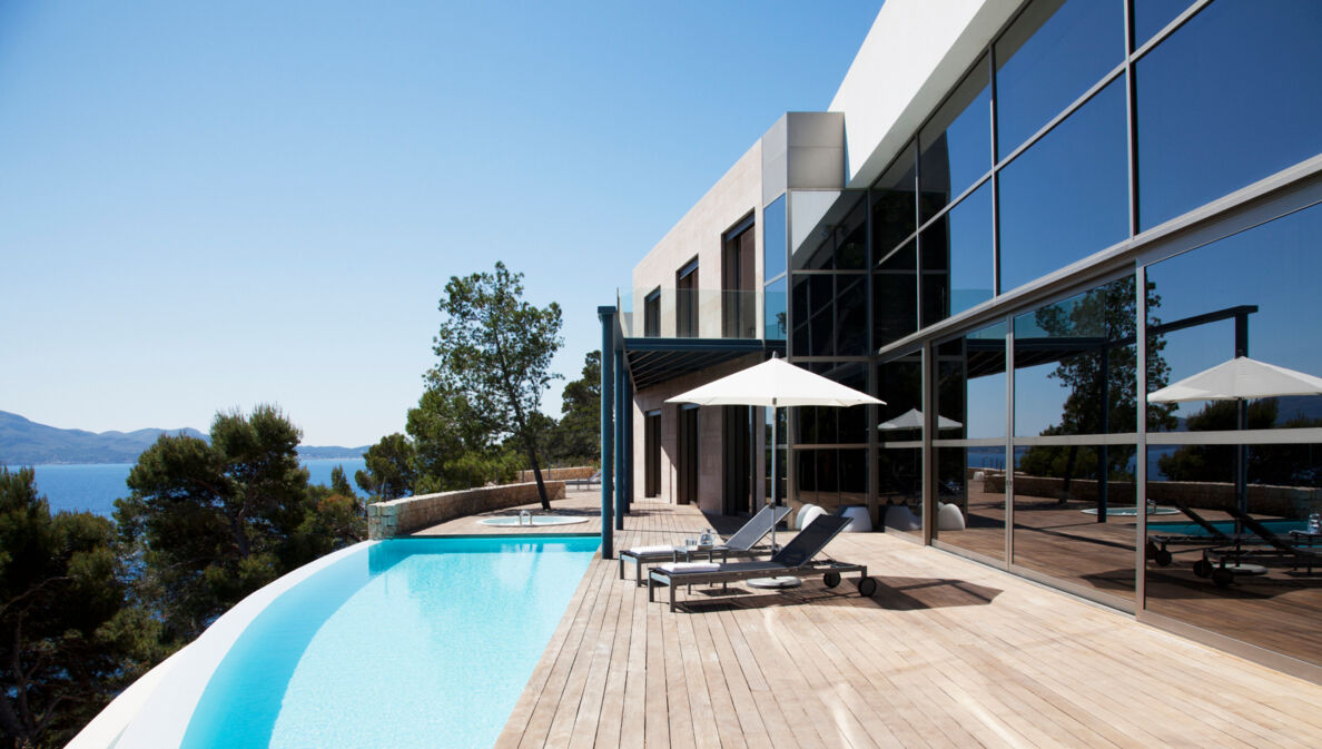 Ein Haus mit großer Glasfensterfront und angeschlossener Holzterrasse mit Pool. Auf der Terrasse befinden sich ein Sonnenschirm und zwei Liegen.