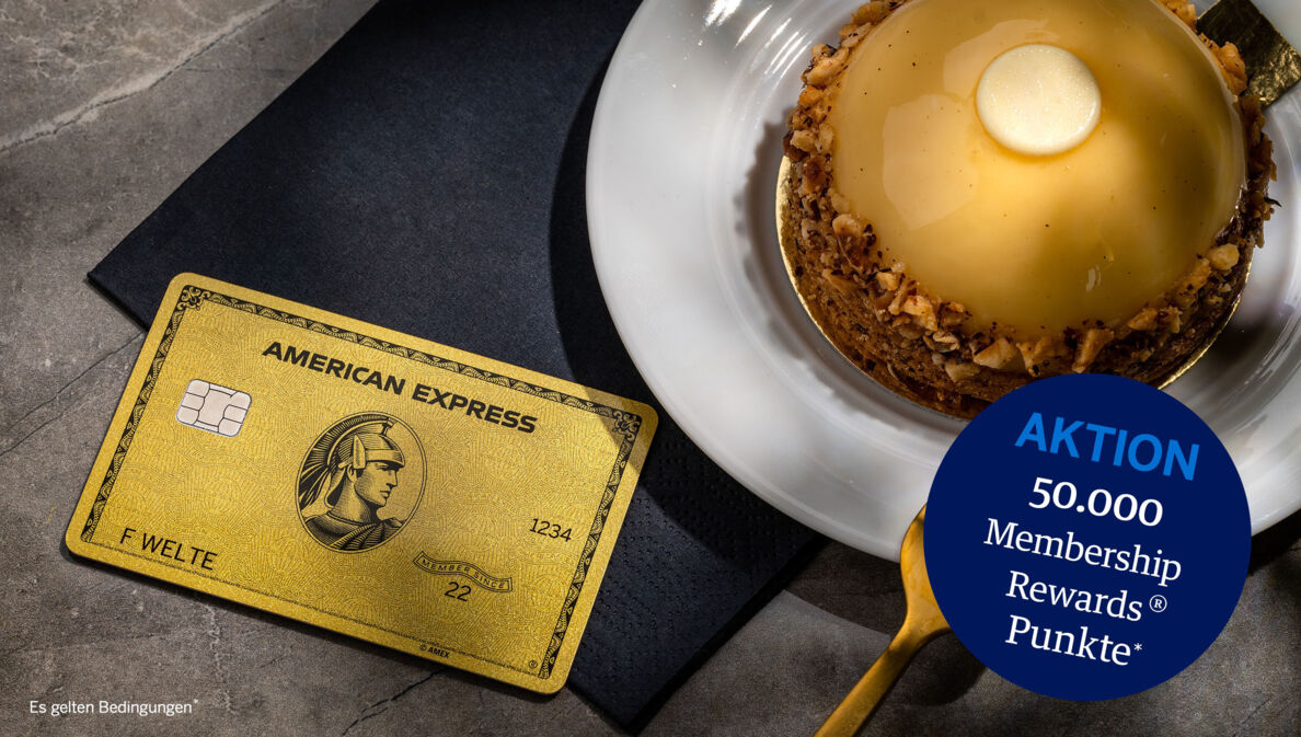 Eine goldene Kreditkarte von American Express liegt auf einer schwarzen Serviette neben einem goldfarbenen Törtchen auf einem Teller und einer goldenen Gabel.