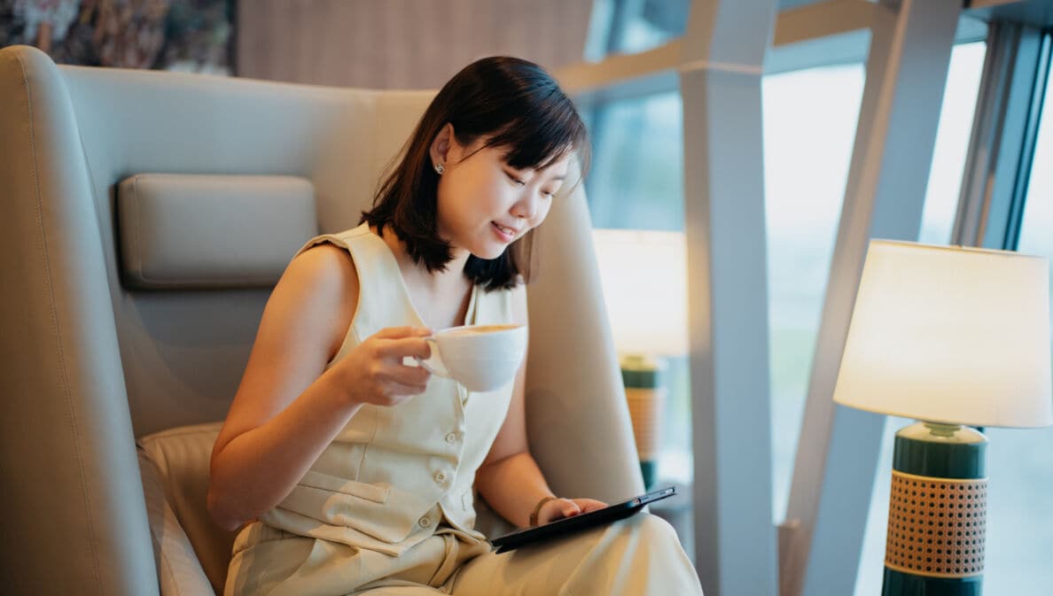 Eine Geschäftsfrau sitzt mit einer Tasse Kaffee in einem Sessel in einer Flughafenlounge und schaut auf ein Tablet in ihrer Hand.