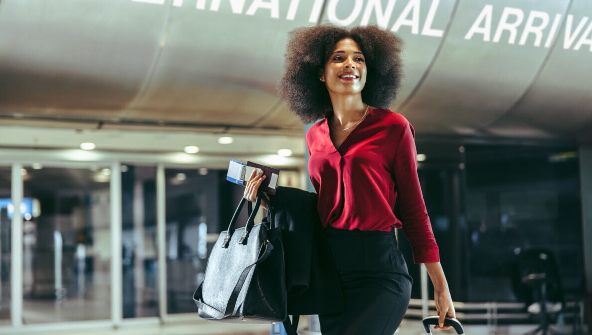 Eine elegante, lächelnde Frau mit Handgepäck vor einem Ausgang am Flughafen.