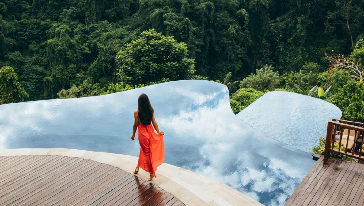 Rückansicht einer Frau im langen Sommerkleid an einem Infinitypool auf einer Terrasse, umgeben von tropischem Regenwald.