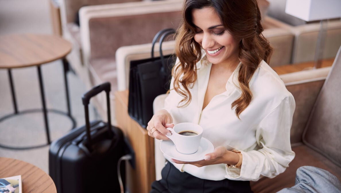 Eine elegant gekleidete Frau sitzt mit einer Tasse Kaffee in einem Sessel, neben ihr steht Gepäck.
