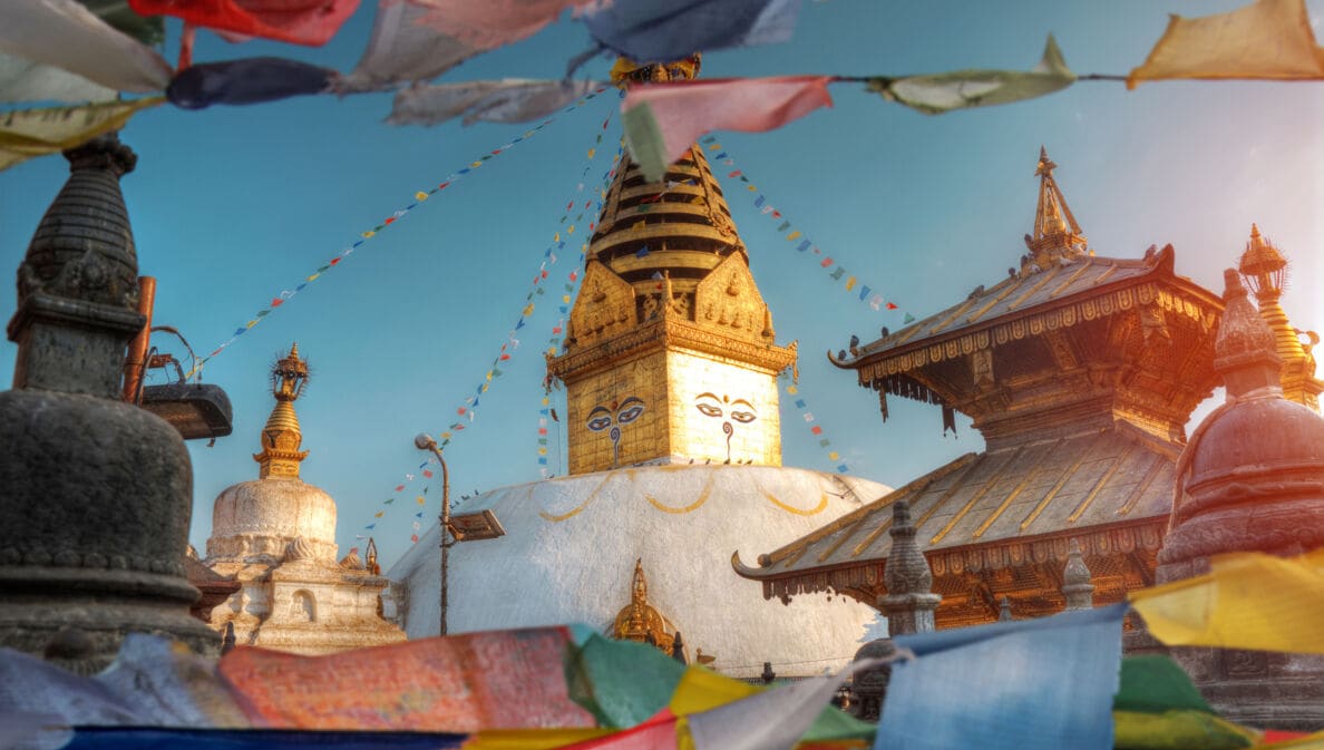 Goldener Turm einer buddhistische Tempelanlage mit aufgemalten Gesichtern neben einer Pagode, im Vordergrund wehende, bunte Gebetsflaggen.