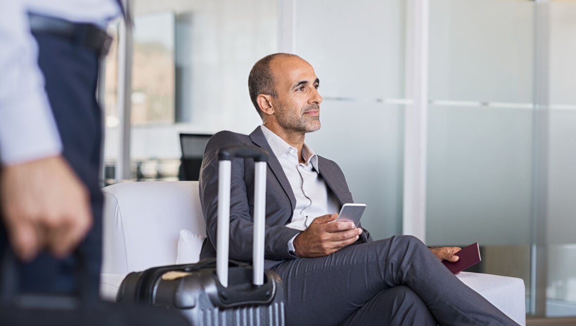 Ein Geschäftsmann sitzt in einem Sessel in einer Flughafenlounge, neben ihm steht ein Koffer.