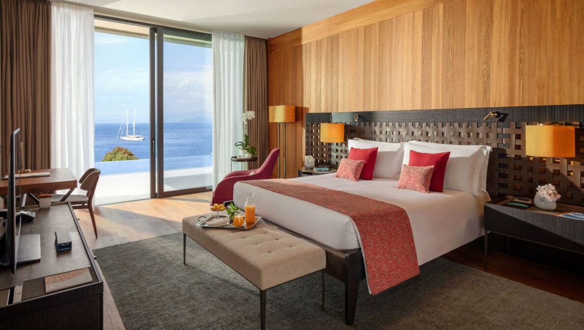 Modern eingerichtete Suite des Hotels Mandarin Oriental Bodrum mit einem großen Bett, einer Sitzbank, Arbeitstisch und raumhohen Fenstern mit Meeresblick.