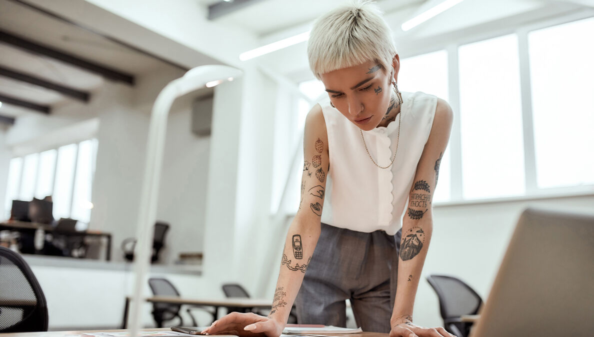 Eine junge tätowierte Frau steht am Schreibtisch und blickt auf verschiedene Entwürfe.
