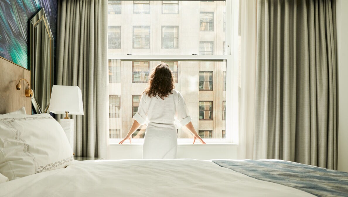 Rückansicht einer Frau im Bademantel, die am Fenster eines Hotelzimmers steht.