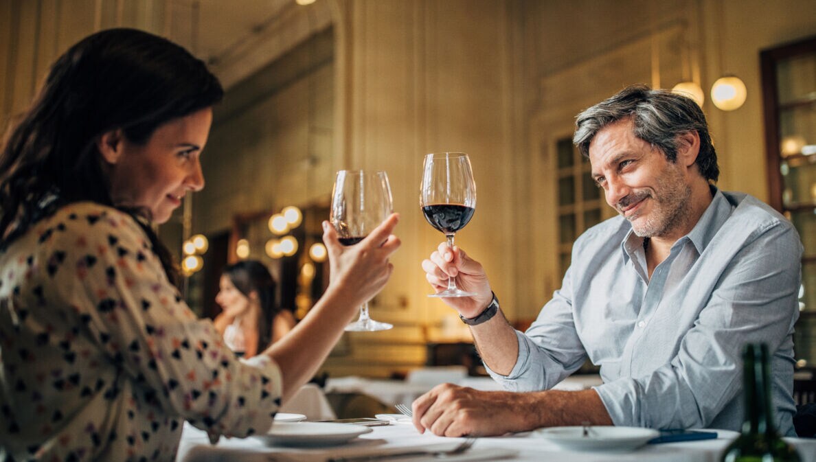 Eine Frau und ein Mann sitzen in einem Restaurant am Tisch, heben je ein Weinglas hoch und lächeln sich an.